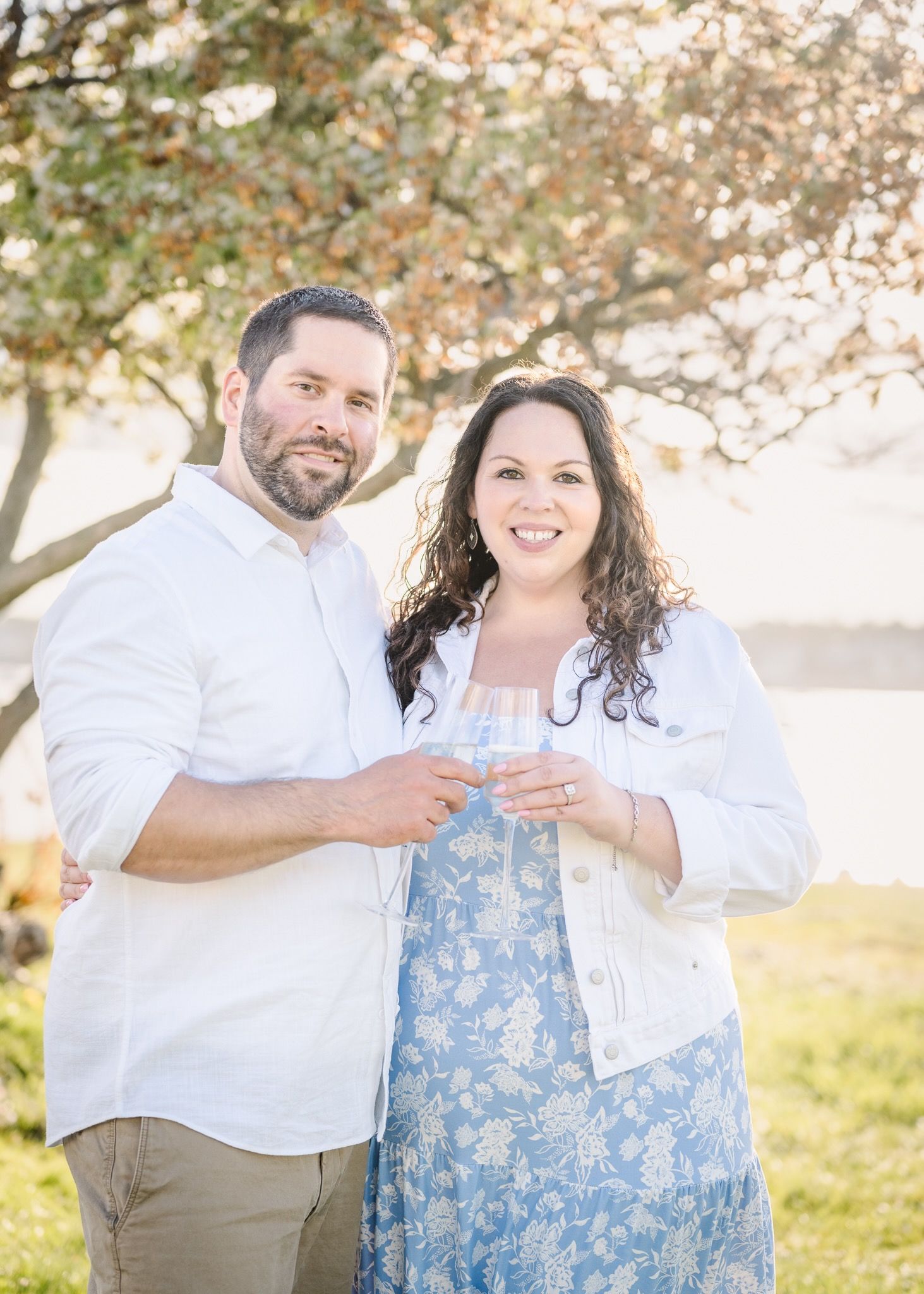A couple in white and blue attire shares drinks while smiling under a flowering tree.