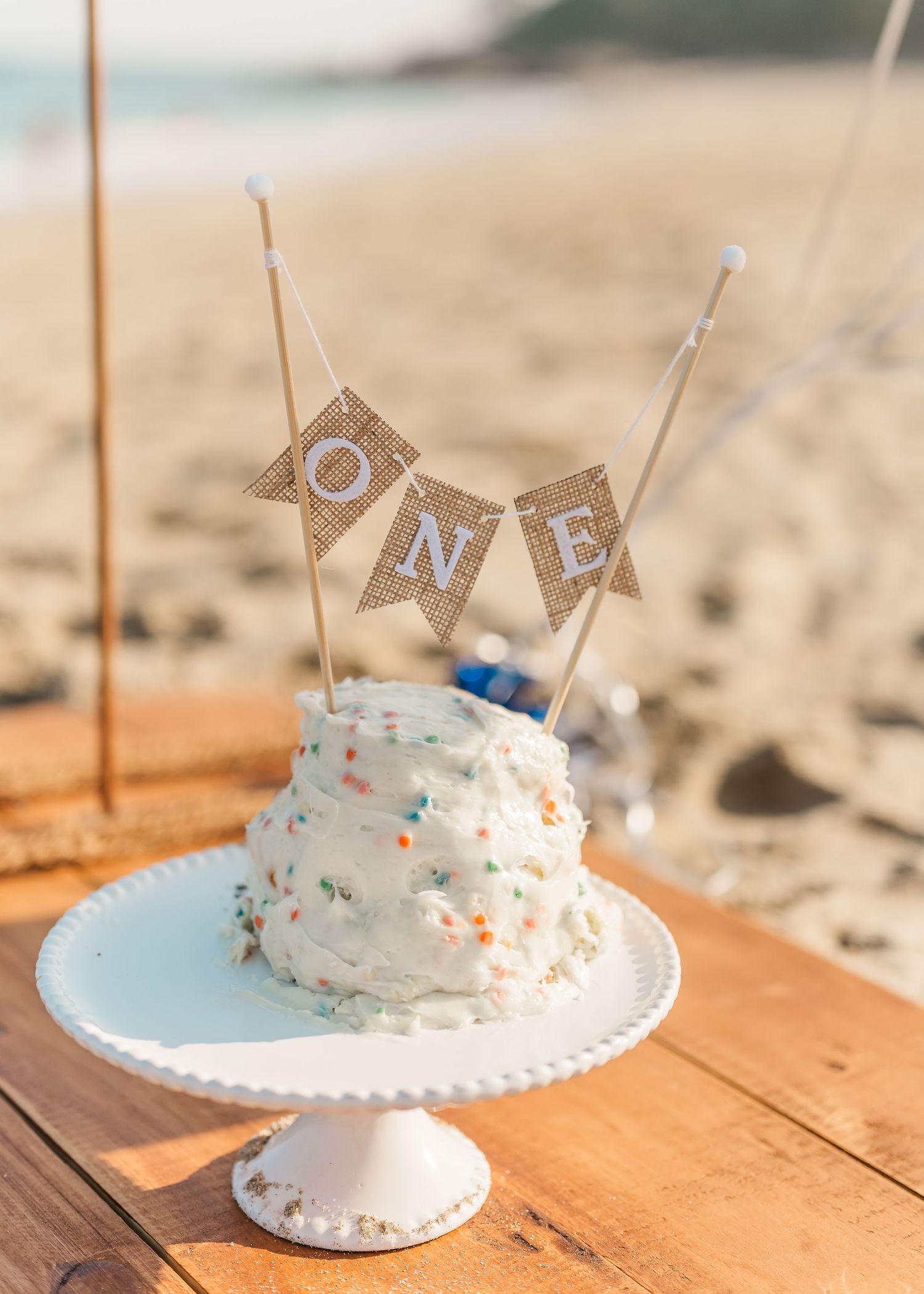 A white smash cake with sprinkles and rustic burlap 'ONE' banner topper sits on a cake stand at the beach.