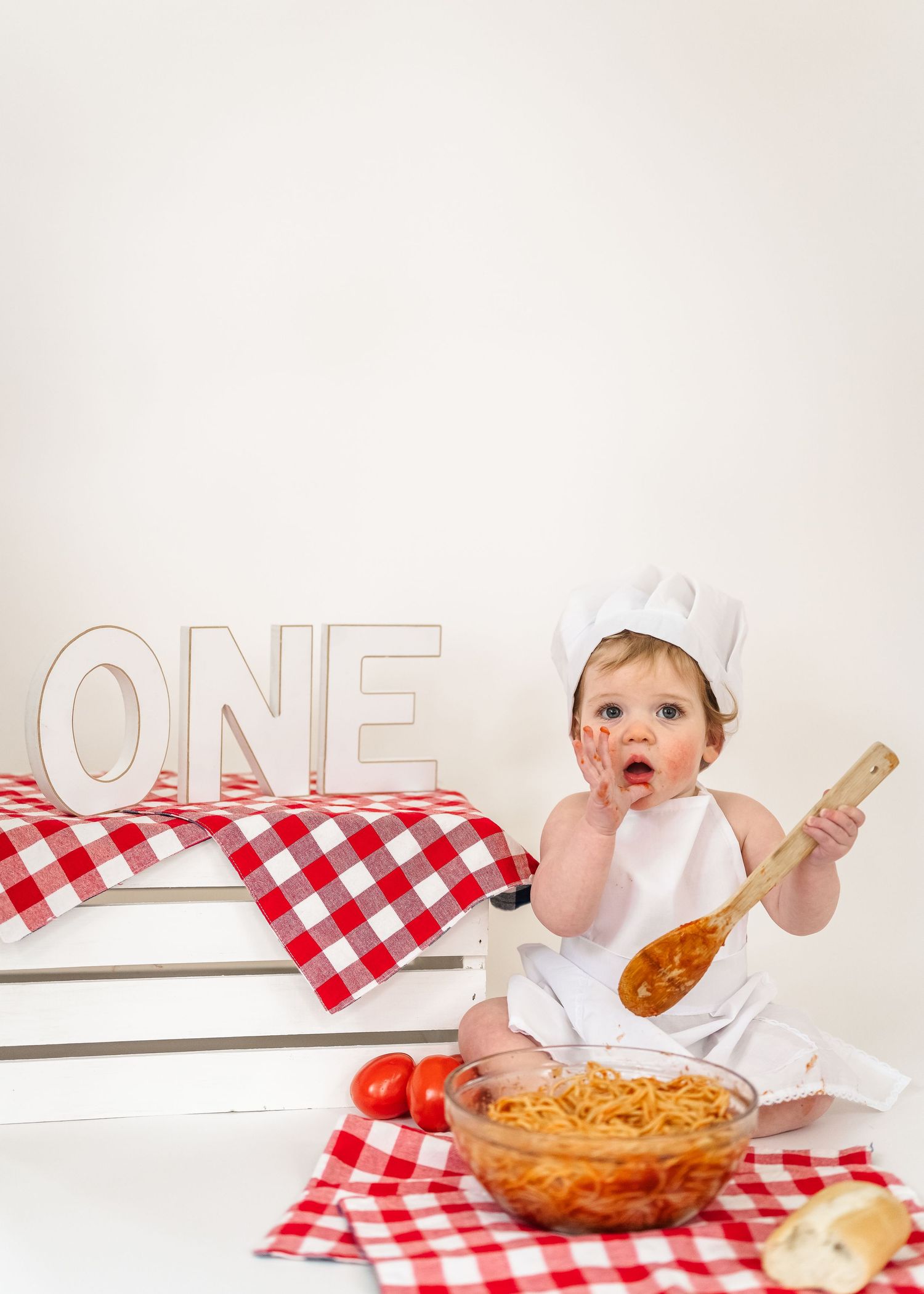 Spaghetti Smash birthday. Baby in chef outfit sits with spaghetti bowl and wooden spoon on red checkered tablecloth next to 'ONE' letters.
