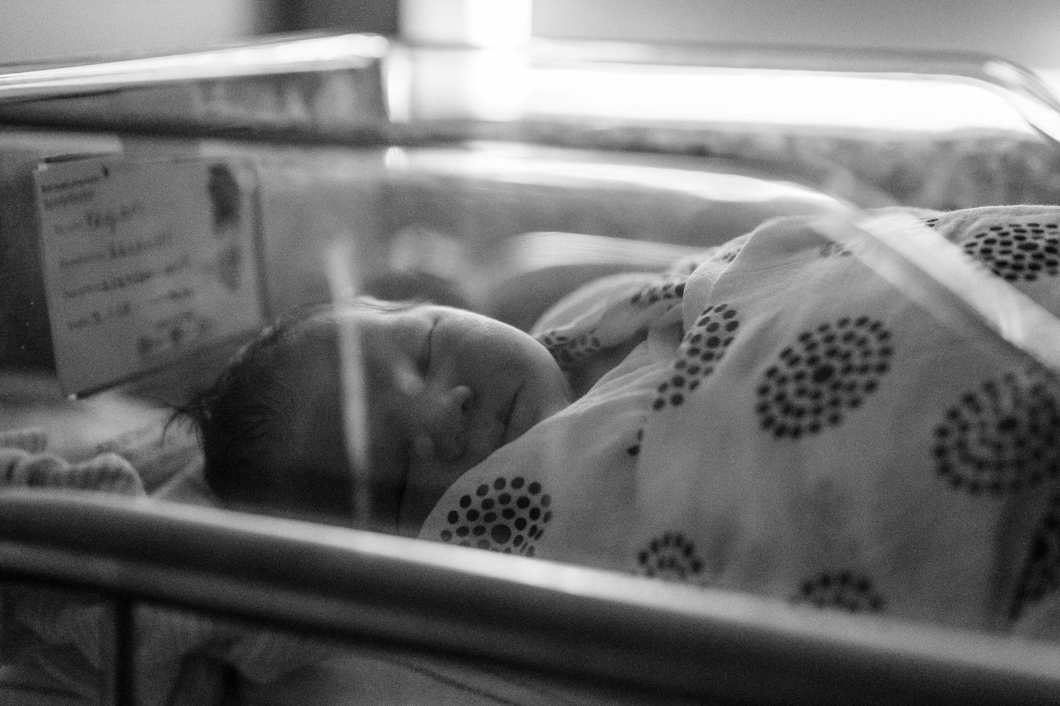 A sleeping newborn baby rests peacefully in a hospital bassinet, wrapped in a blanket with polka dot patterns.