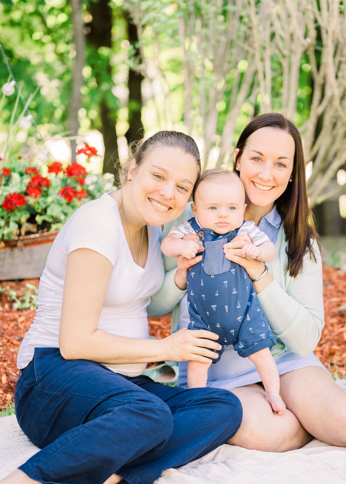 Family poses together on blanket with baby in blue romper against garden backdrop.