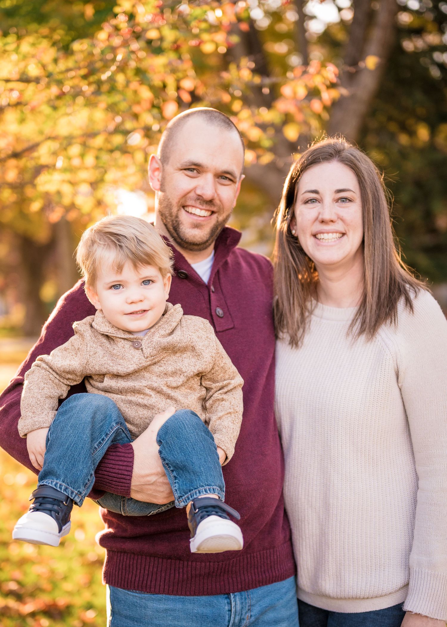Outdoor family portrait with autumn trees and golden sunlight in background.