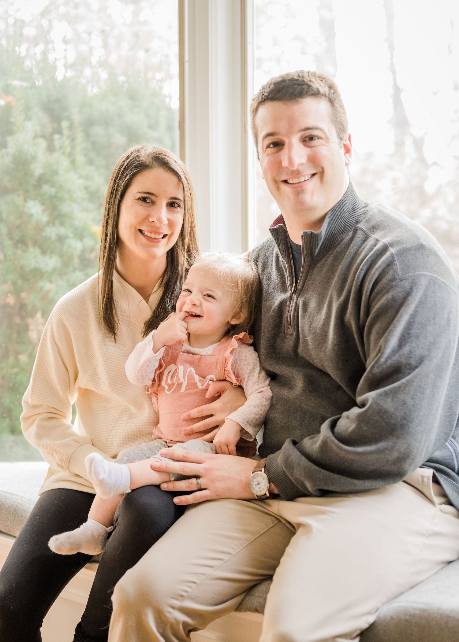Family of three poses together on couch by window for holiday portrait.