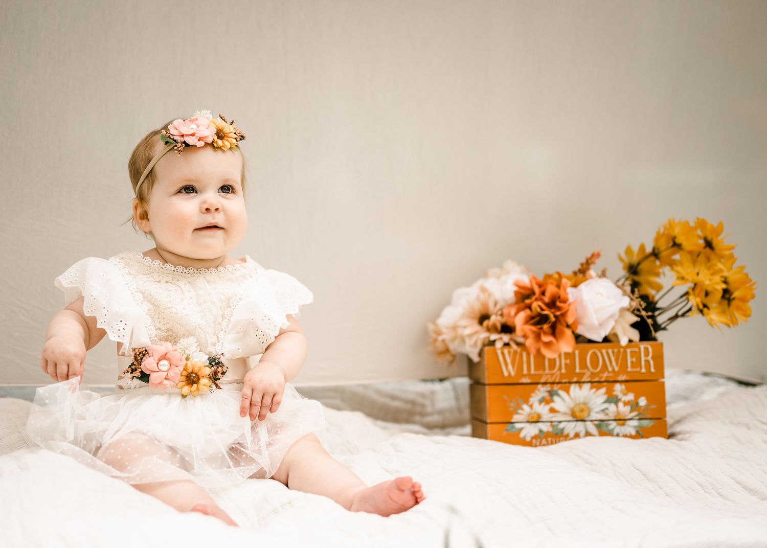 Baby in white lace dress sits on white sheets with floral decorations and wooden crate.