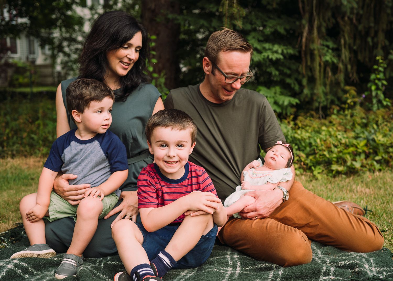 Family relaxing together on blanket in backyard with newborn baby.