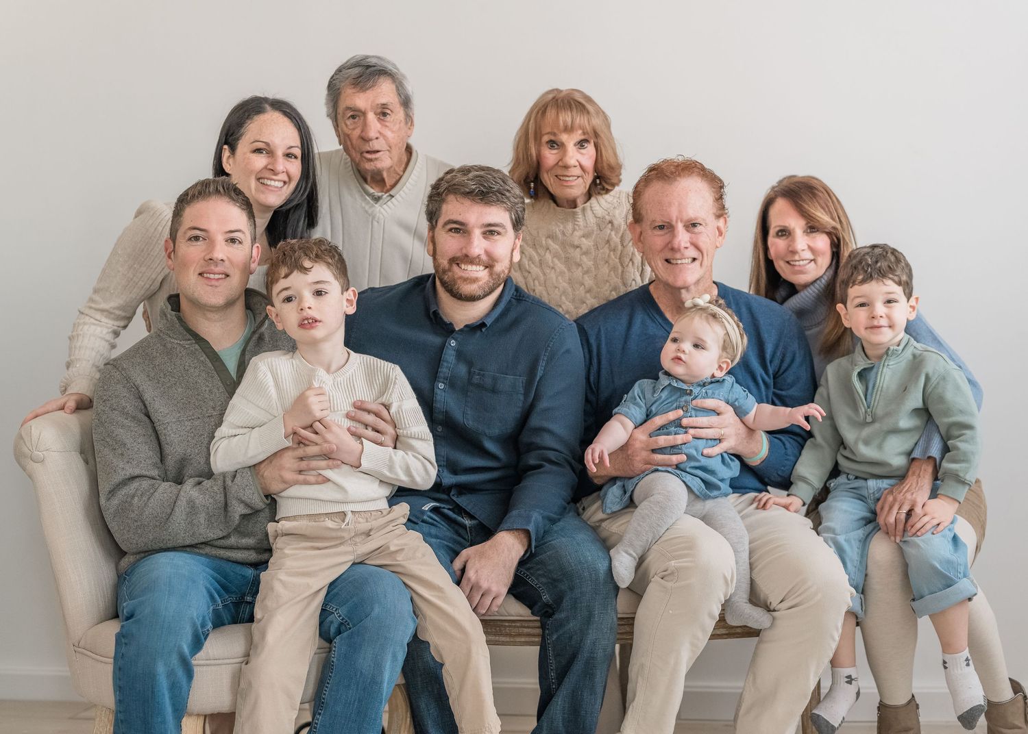 Multi-generational family wearing coordinated neutral colors poses together on a light colored couch for a portrait.