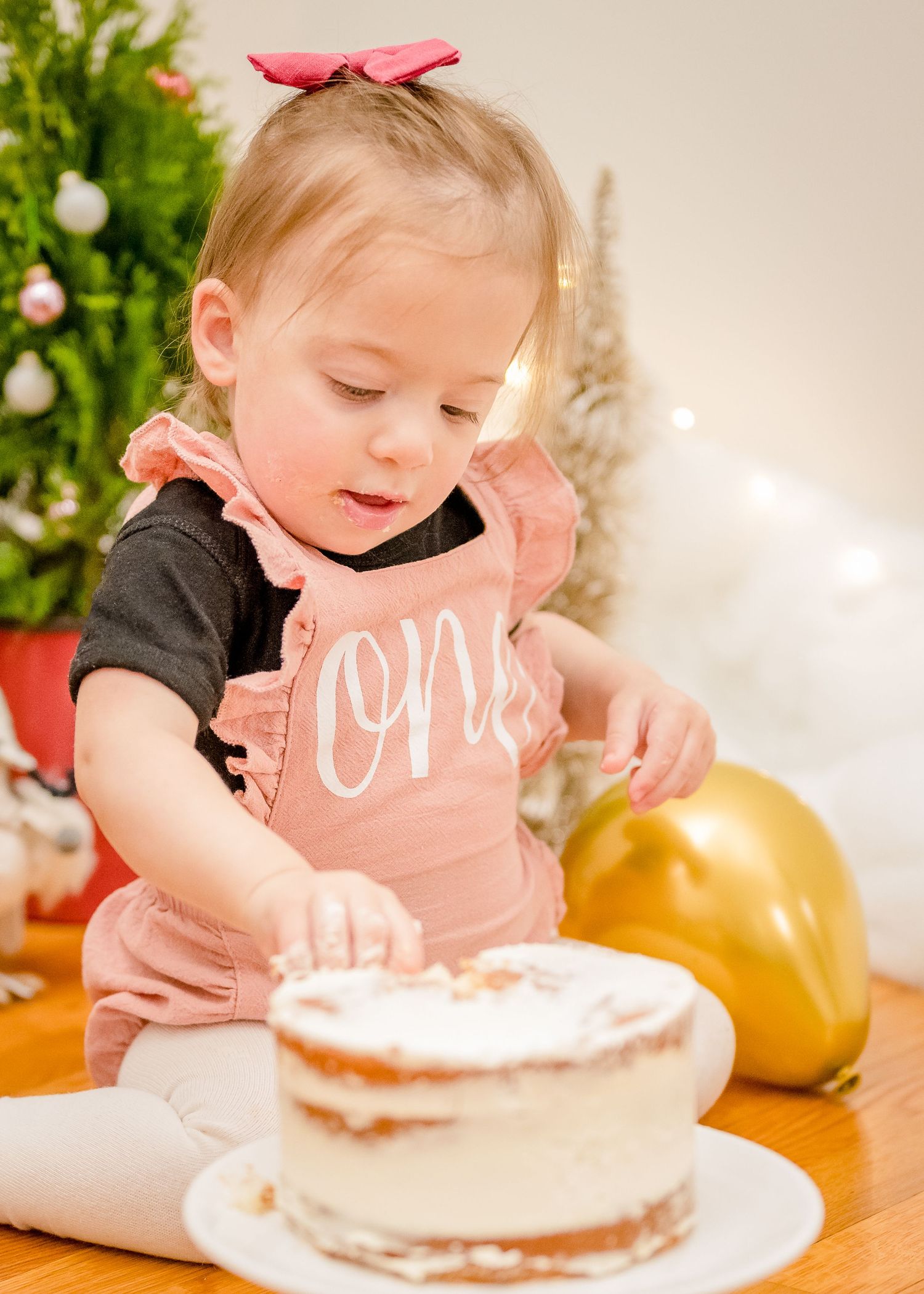 Small child in pink bib explores a white cake with holiday decorations in the background.