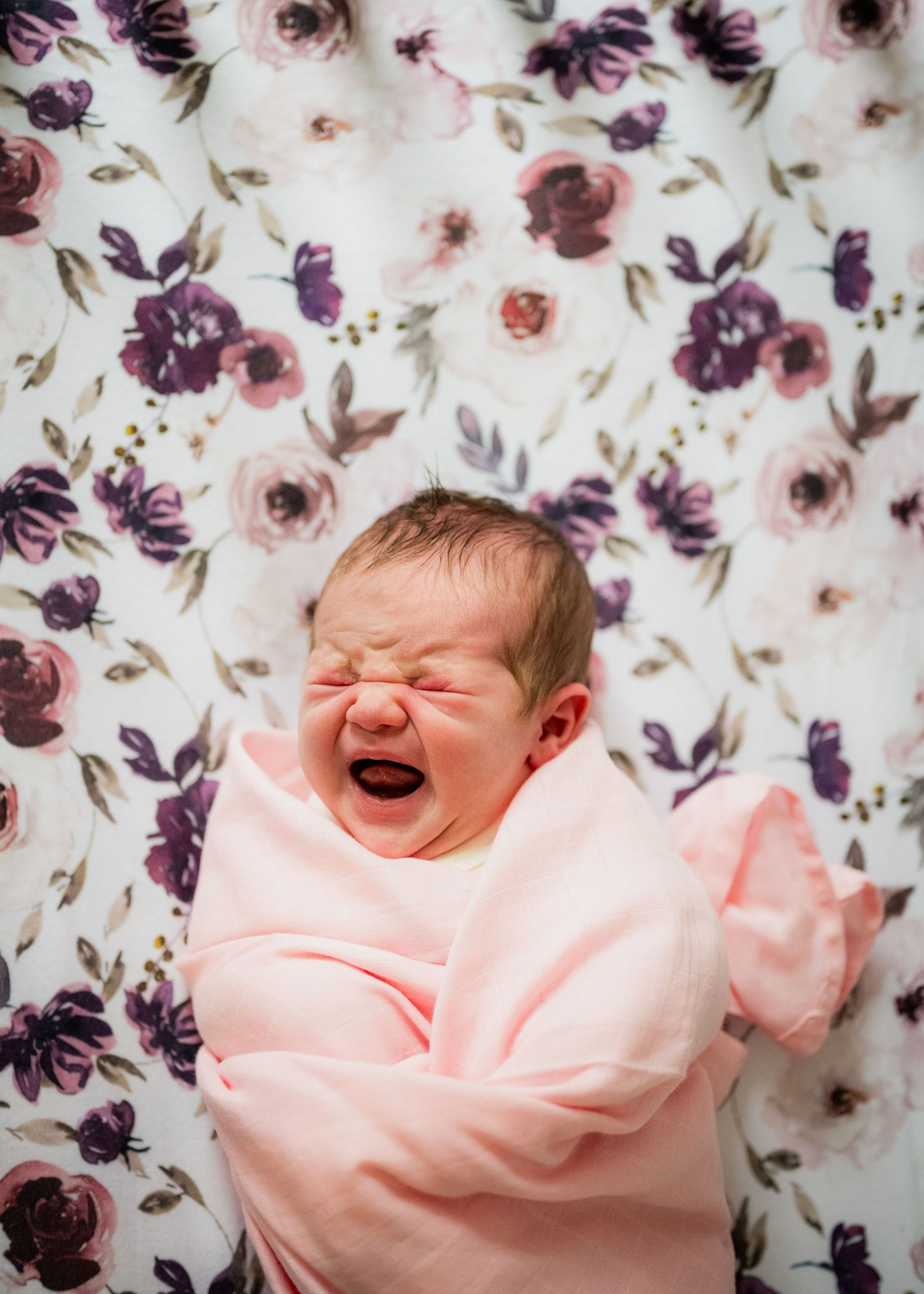 Laughing baby wrapped in soft pink blanket lying on floral pattern background with purple and burgundy flowers.