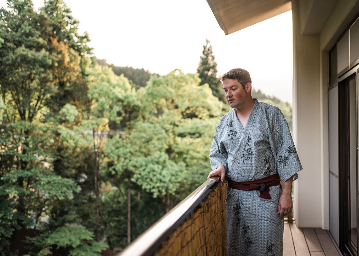 Person in traditional Japanese yukata standing on wooden balcony overlooking forest.