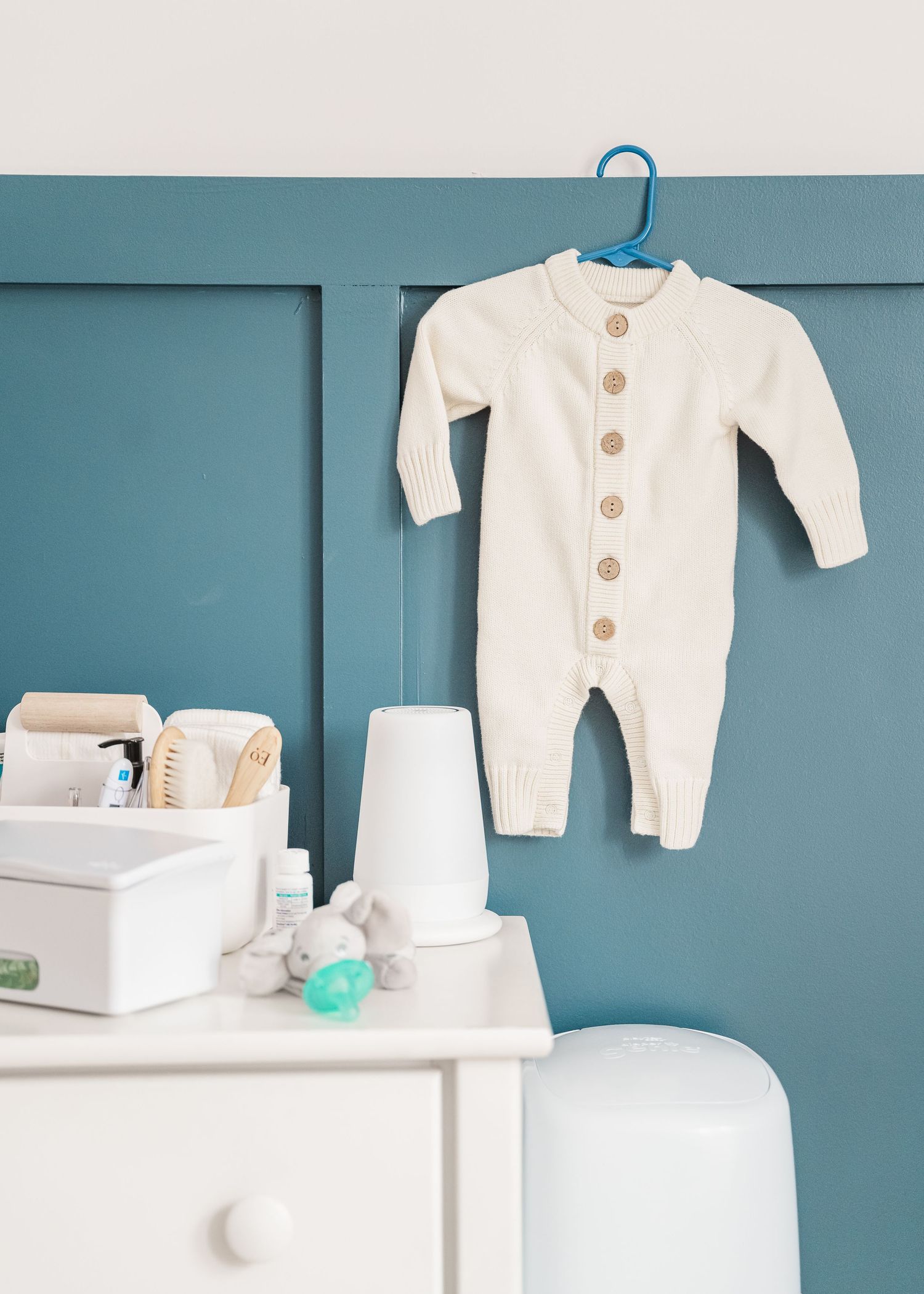 White baby onesie hanging on blue wall with nursery essentials displayed on dresser.