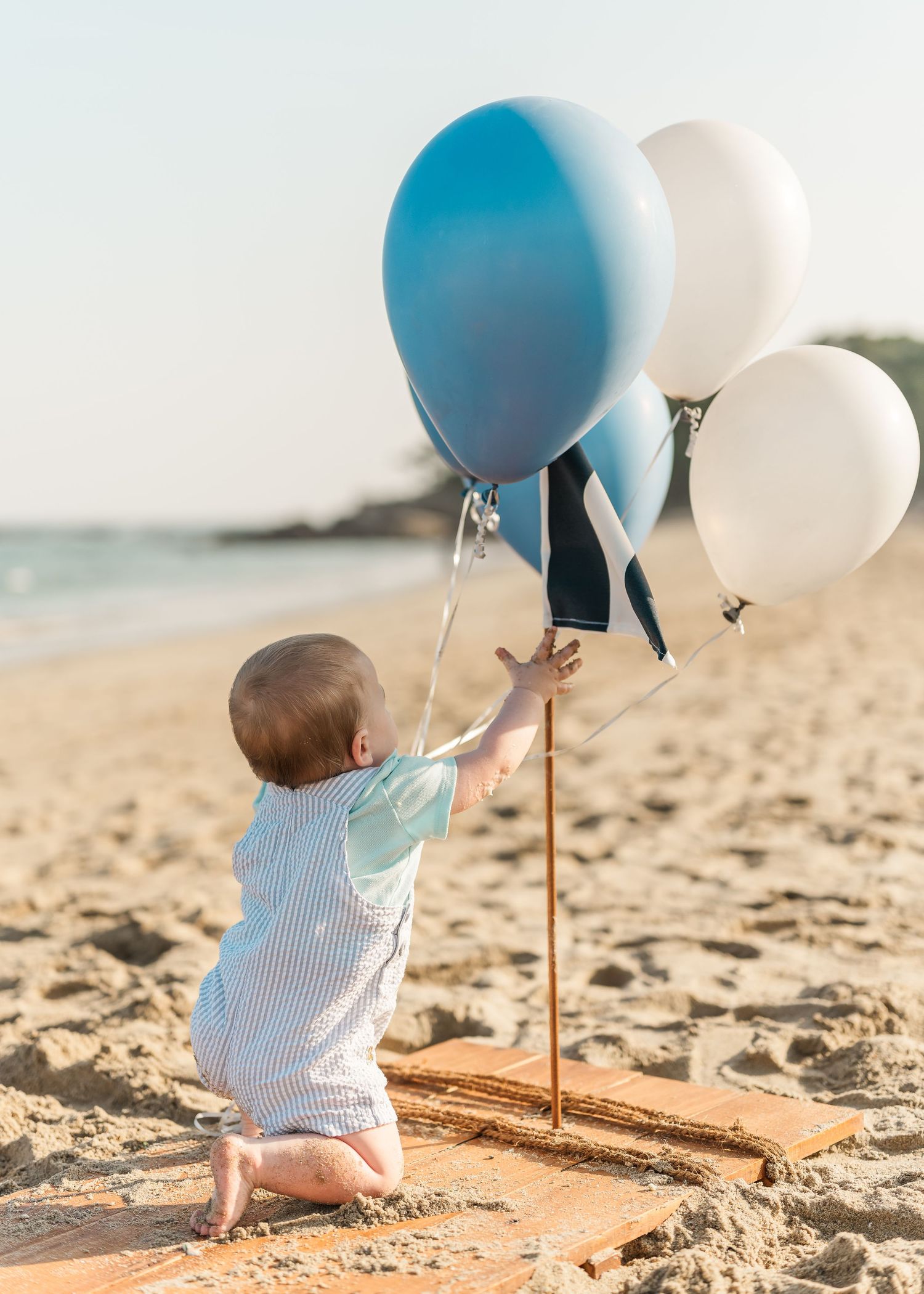 A baby in striped onesie sits on wooden platform at beach with blue and white balloons during sunset.