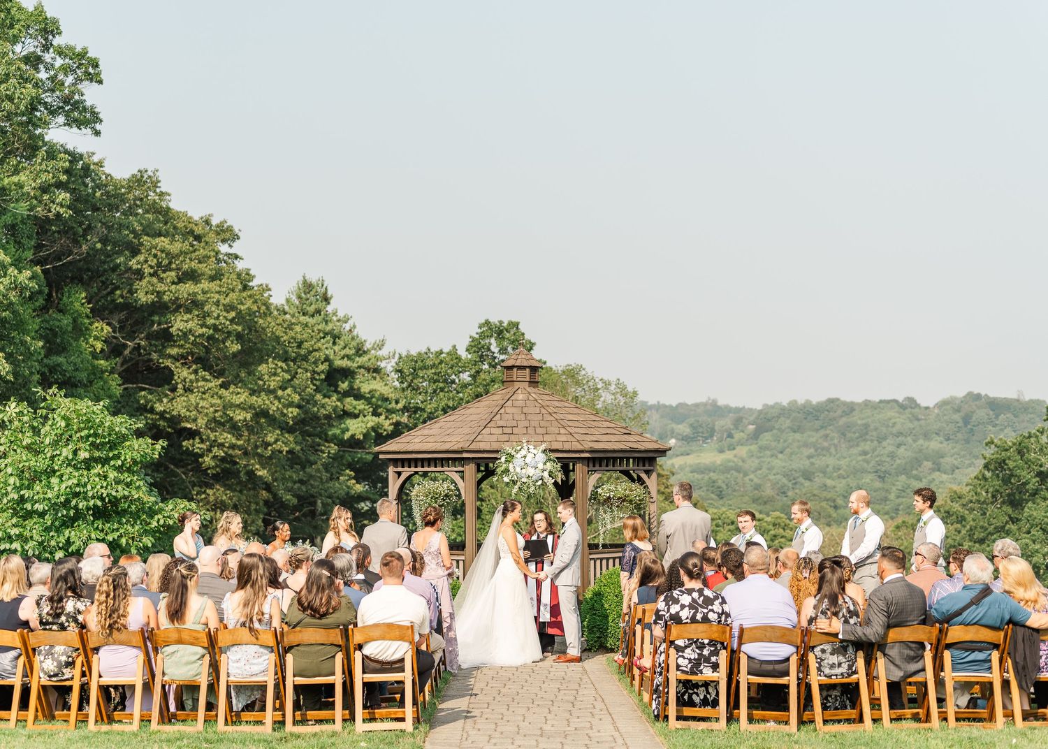 Wedding guests seated on wooden chairs in an outdoor garden ceremony venue with lush greenery and a gazebo.