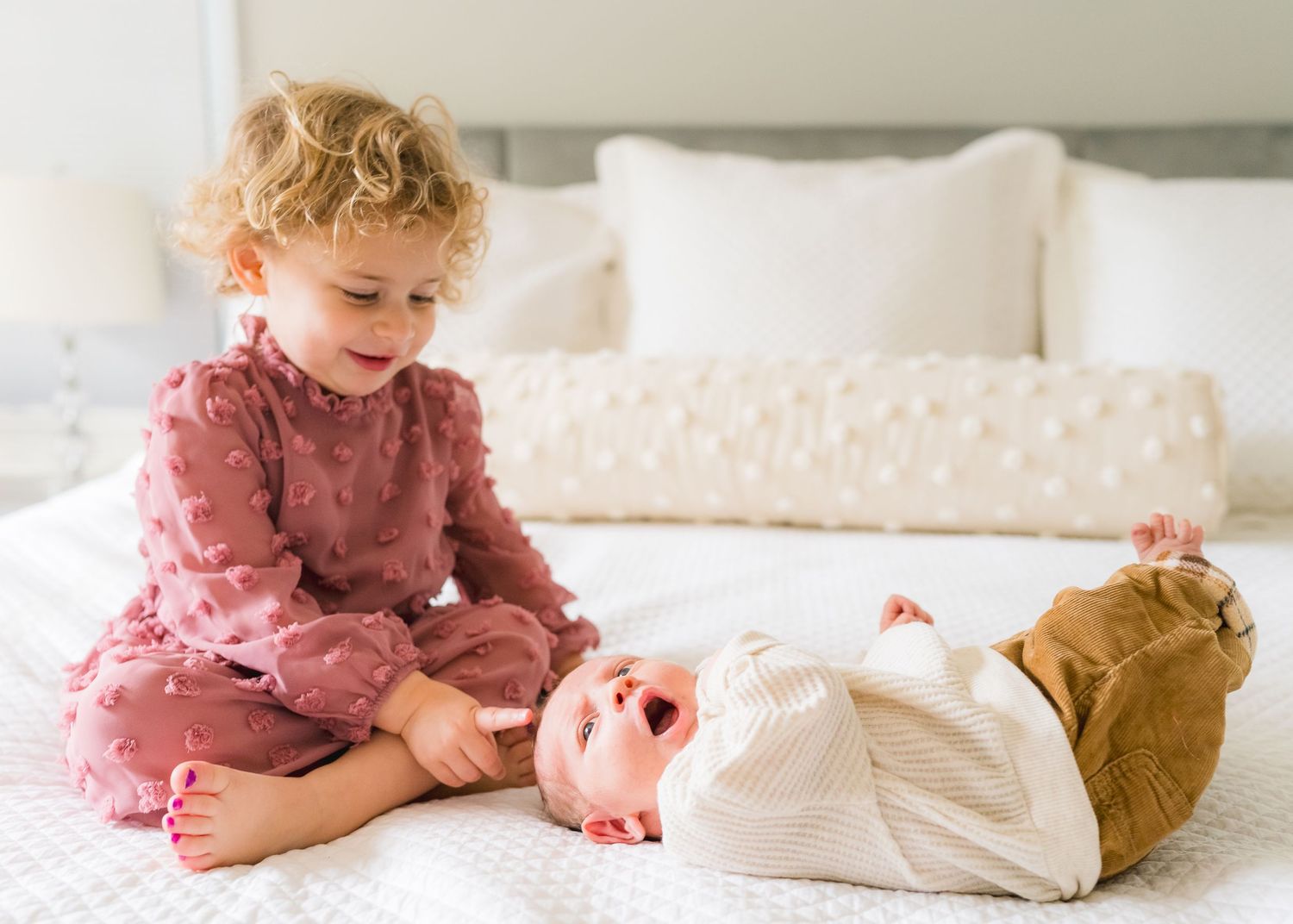 Toddler in pink dress playing with baby sibling lying on white bedding.