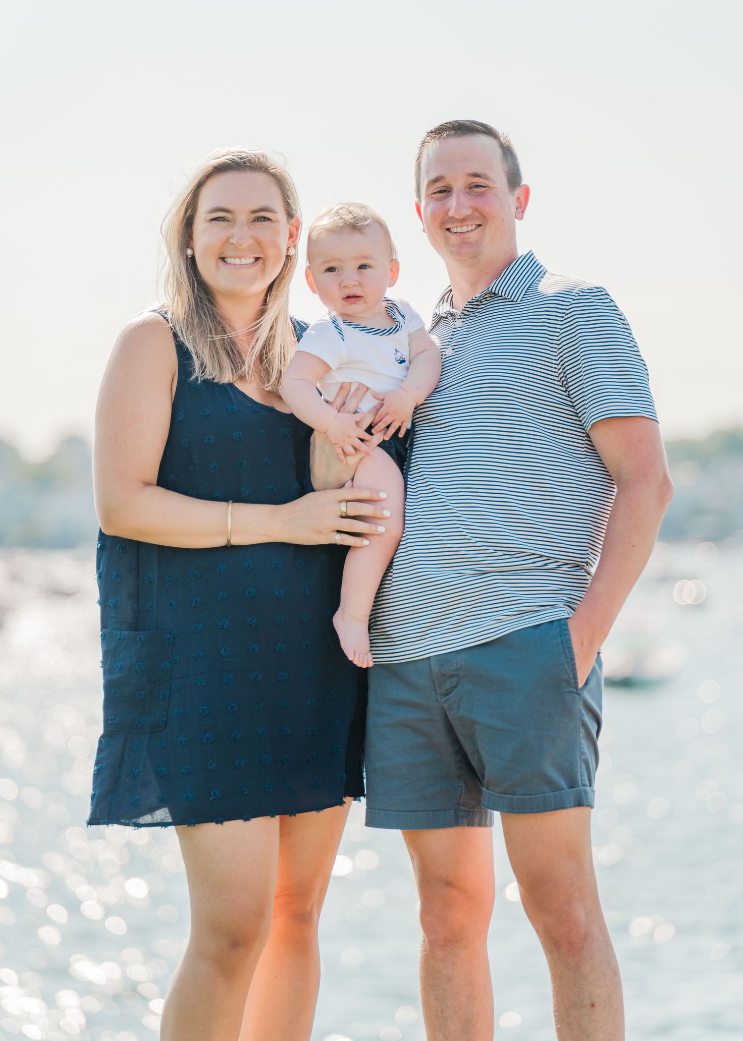 A happy family of three poses together on a bright sunny beach with sparkling water in the background.