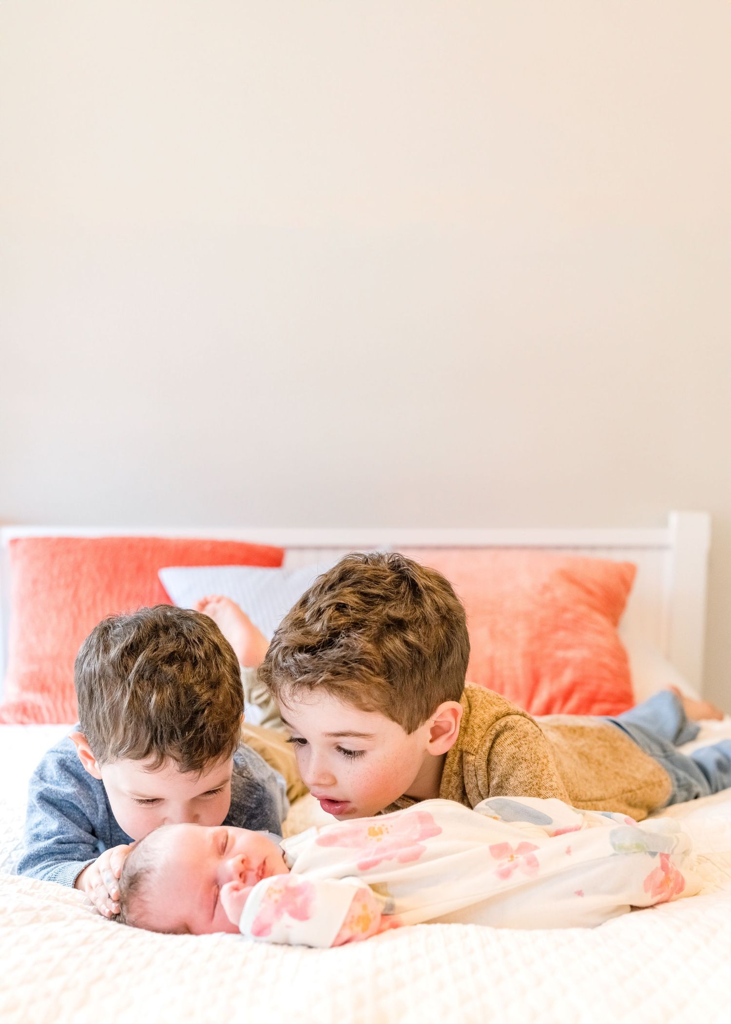 Two boys lie on a bed with soft coral pillows, looking down with gentle interest.