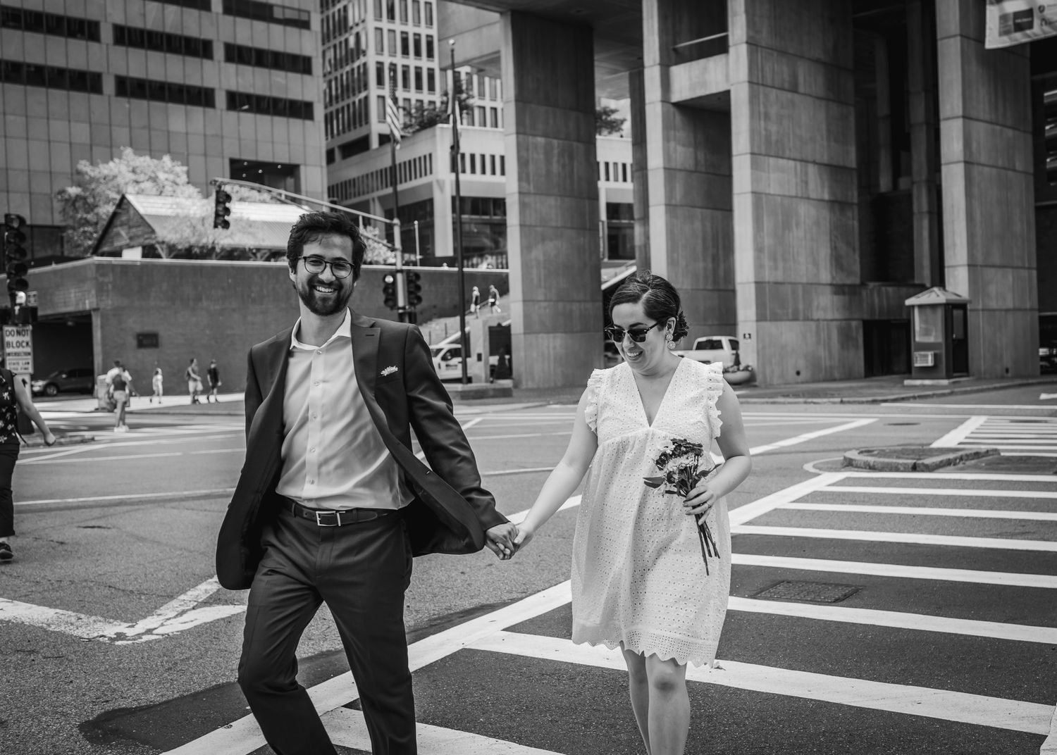 A black and white street photo of a couple walking across an urban intersection.