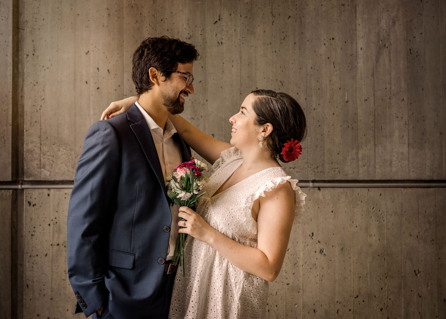 A couple shares a joyful moment against a concrete wall while holding a bouquet of flowers.