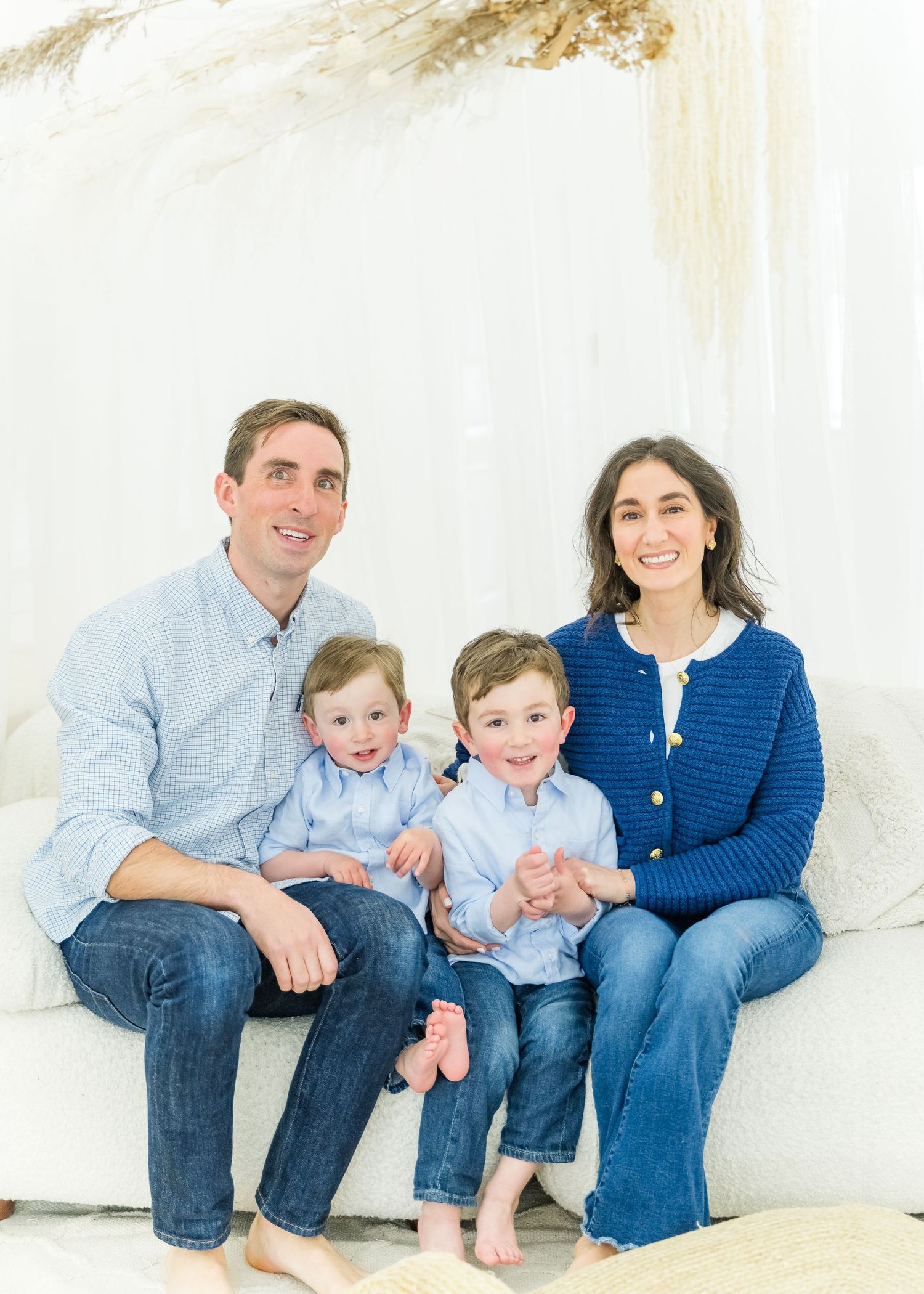 Family of four sits together on white couch wearing coordinating blue and denim outfits for casual portrait.
