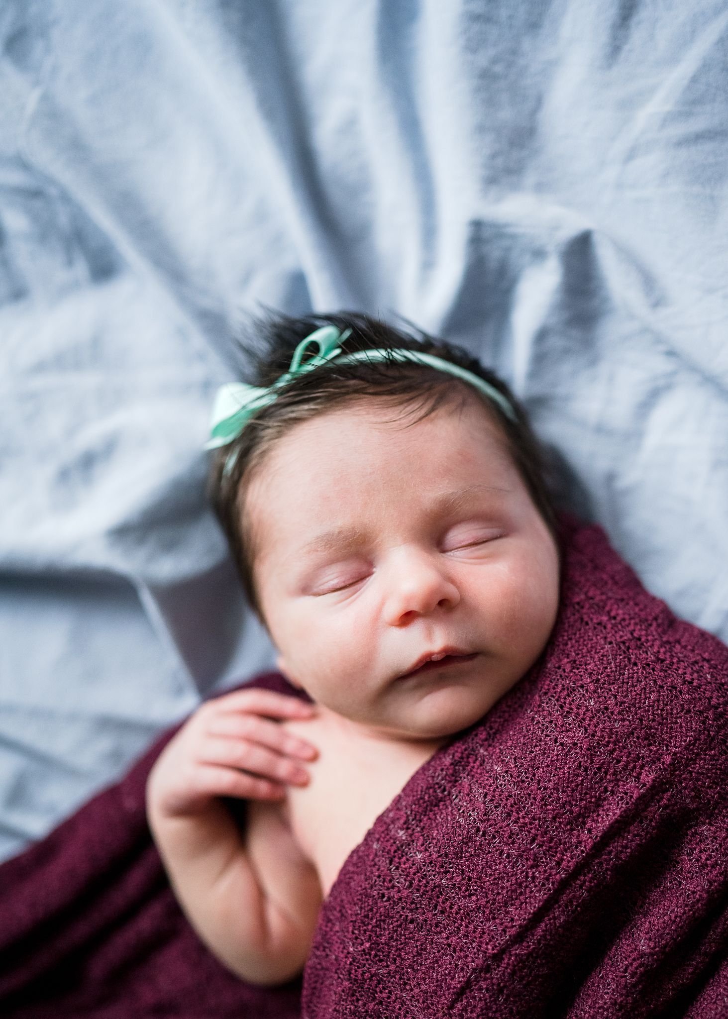 Sleeping newborn with green headband wrapped in burgundy blanket on blue bedding.