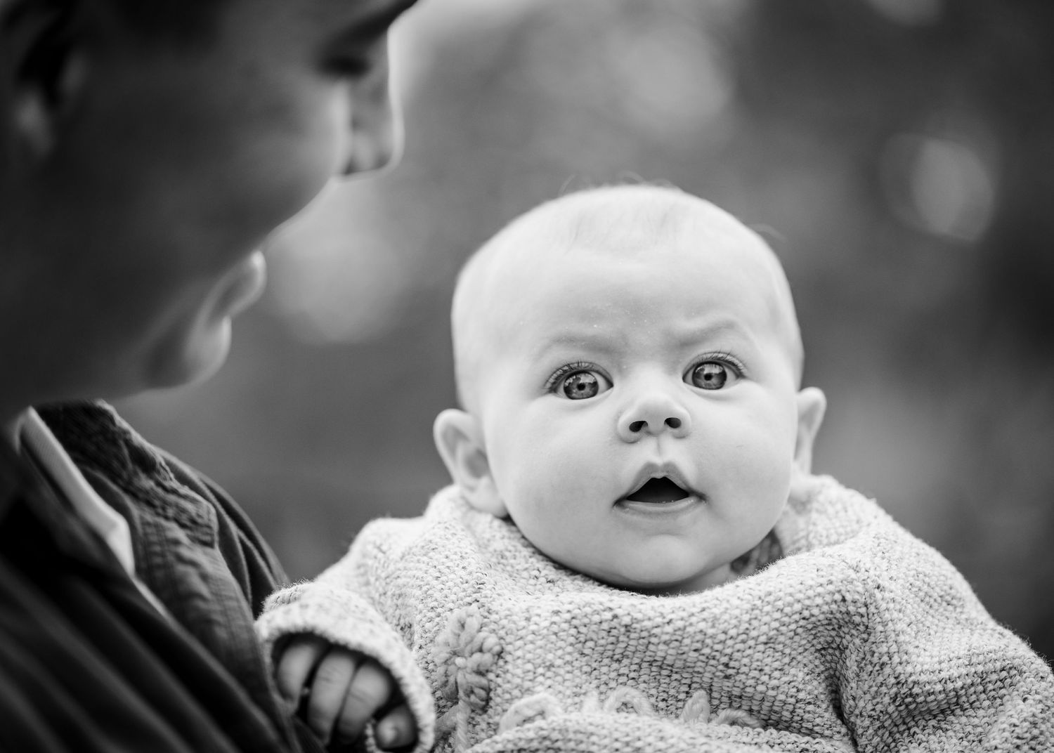 A baby wearing a knit sweater gazes upward with wide-eyed curiosity in black and white portrait.