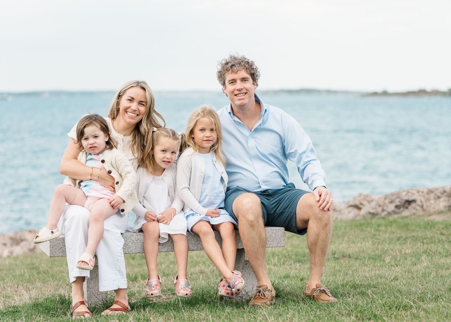 Family sitting together on grassy hill overlooking ocean coastline in casual summer clothing.