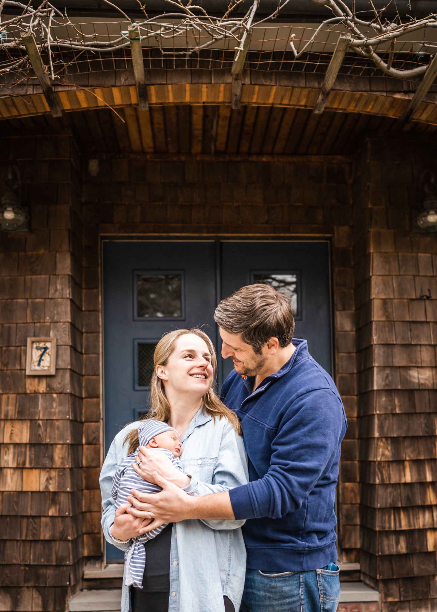 A couple sharing a tender moment while holding their newborn baby outside a rustic wooden house.