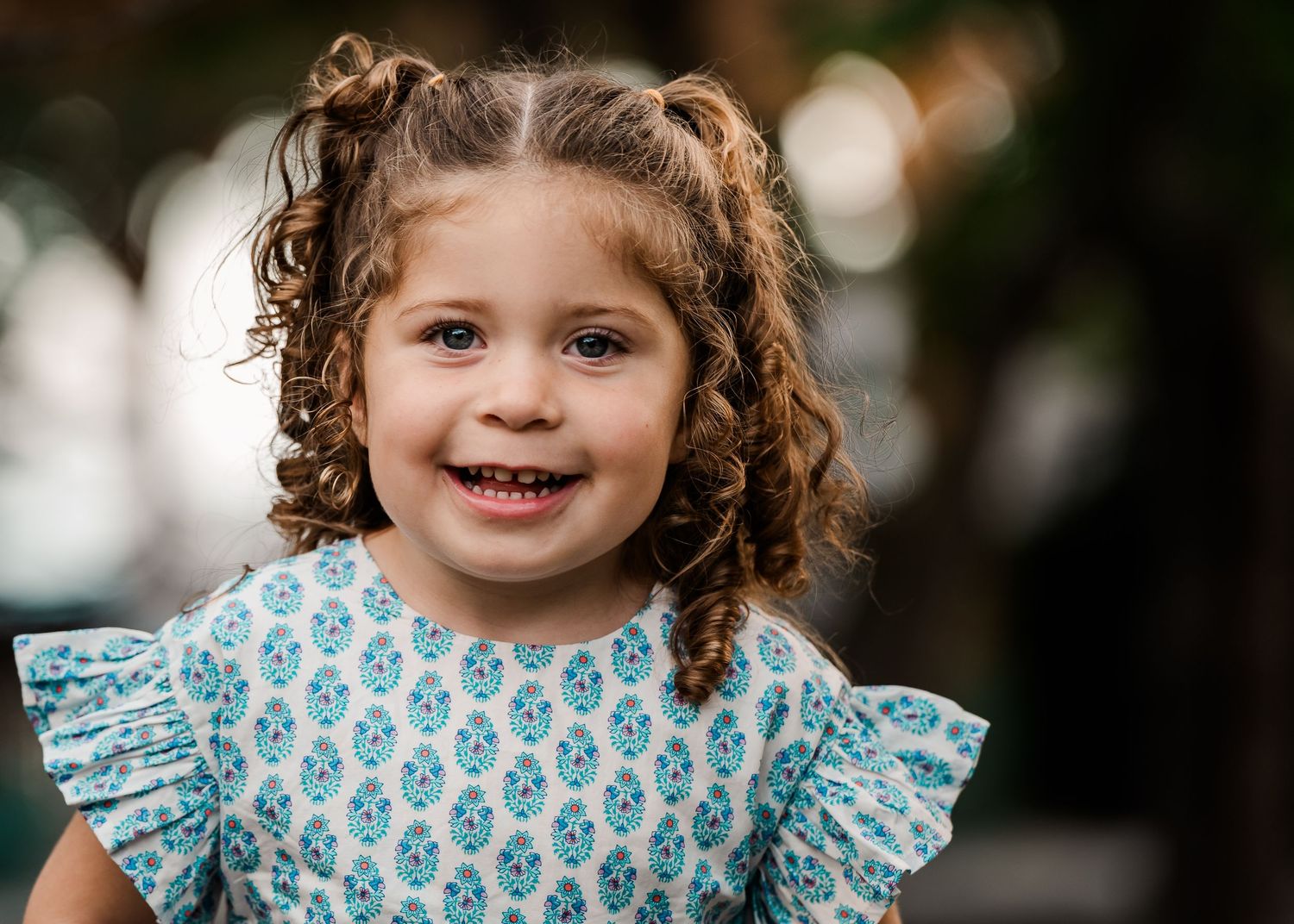 A smiling child wearing a blue patterned dress with ruffled sleeves and curly hair against a blurred background.