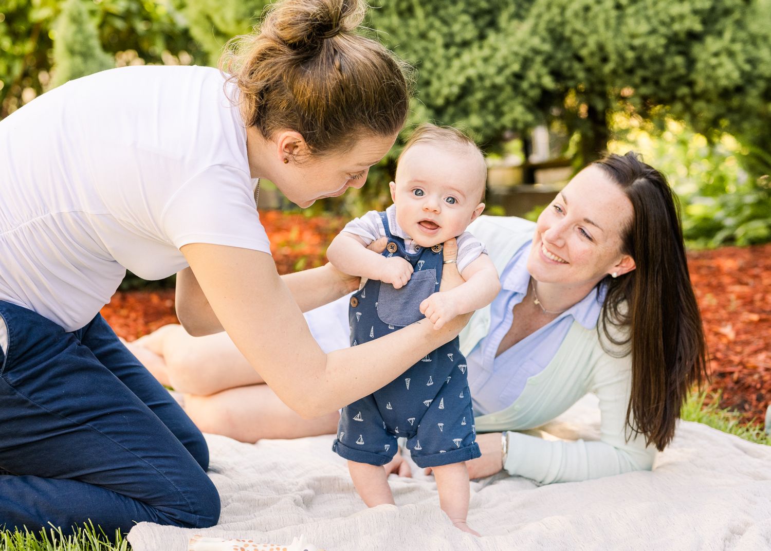Baby in navy outfit playing outdoors on white blanket with greenery in background.