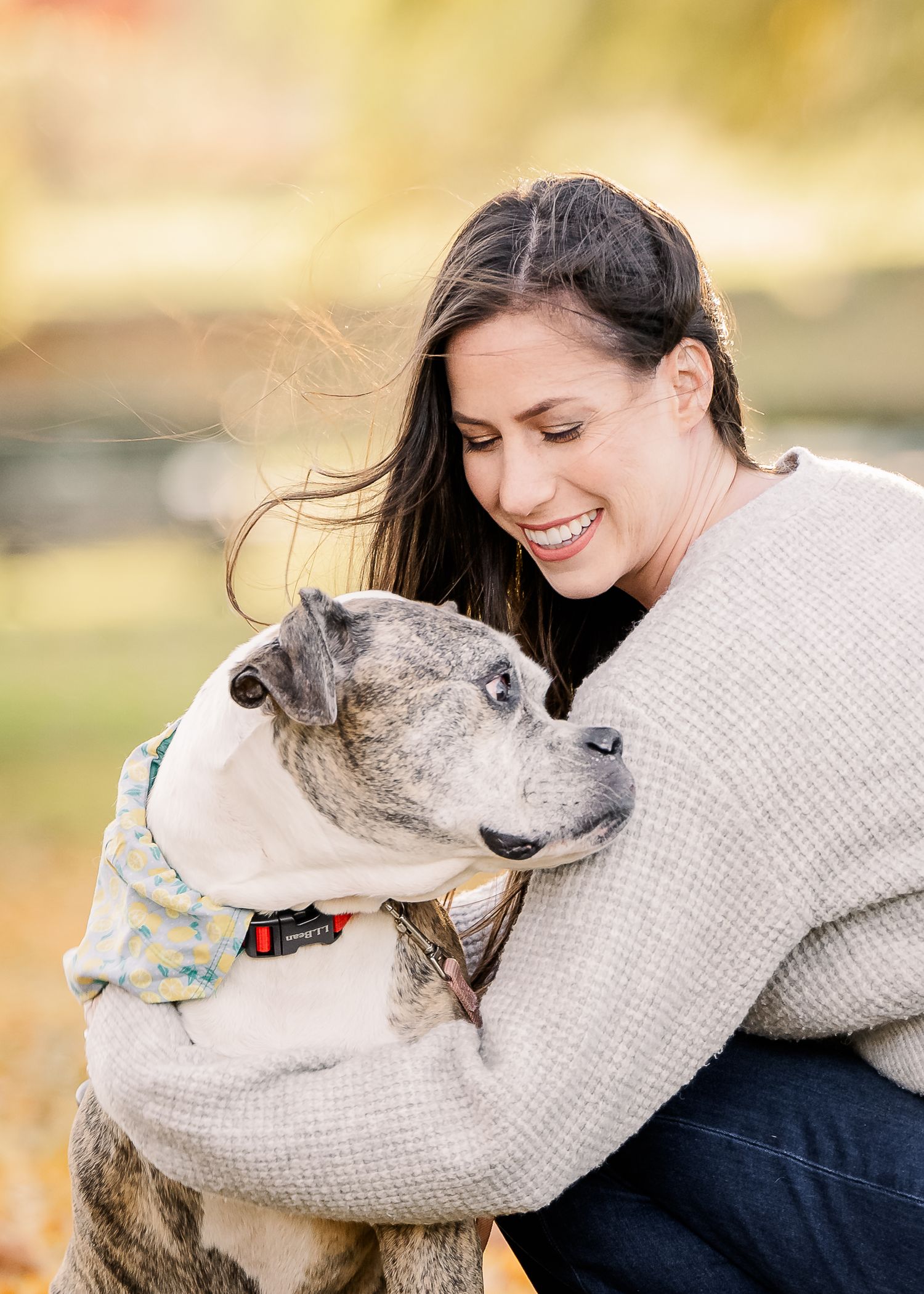 A person in a beige sweater shares a tender moment hugging their gray and white pit bull dog outdoors.