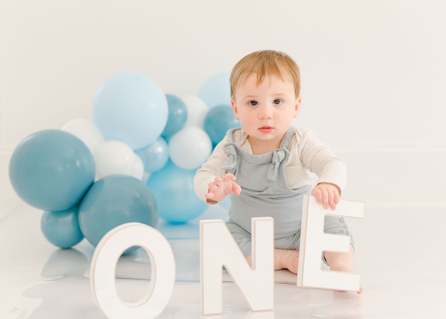 First birthday photo setup with white 'ONE' letters and soft blue and white balloons in the background.