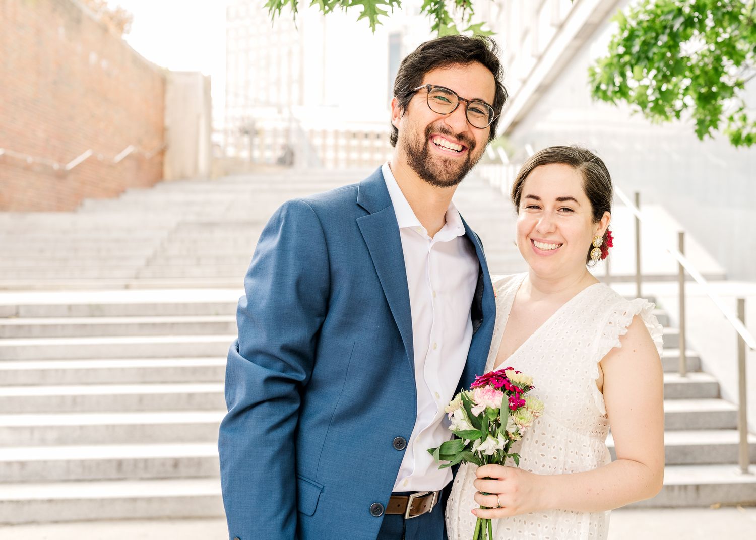 A couple in wedding attire smiles together on stone steps with pink flowers on a sunny day.