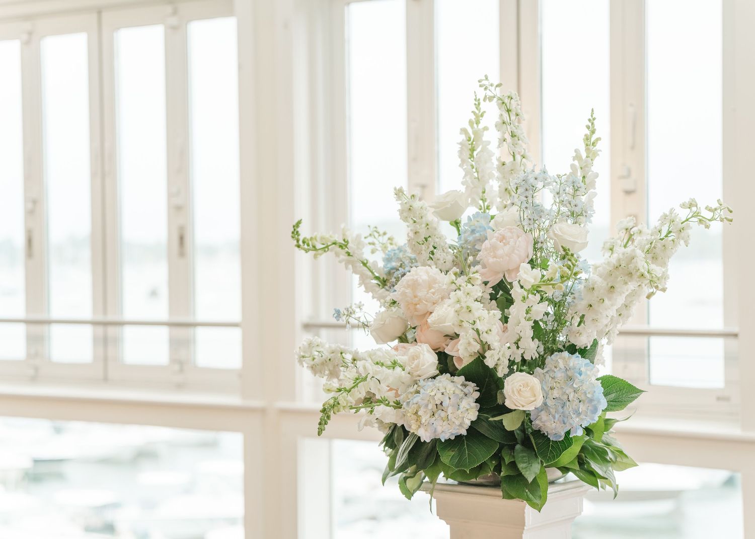 Elegant white and blush floral arrangement with hydrangeas and delphinium in a bright window-lit setting.