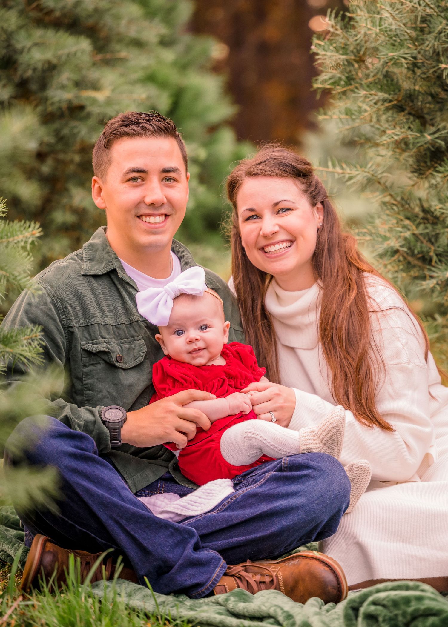 Family portrait outdoors among evergreen trees sitting on blanket during holiday photo session.