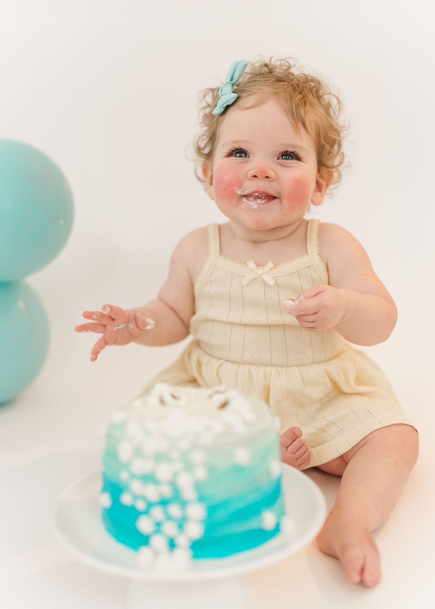 Cake smash. A baby in a cream-colored romper sits excitedly next to a blue ombre cake with white decorations.