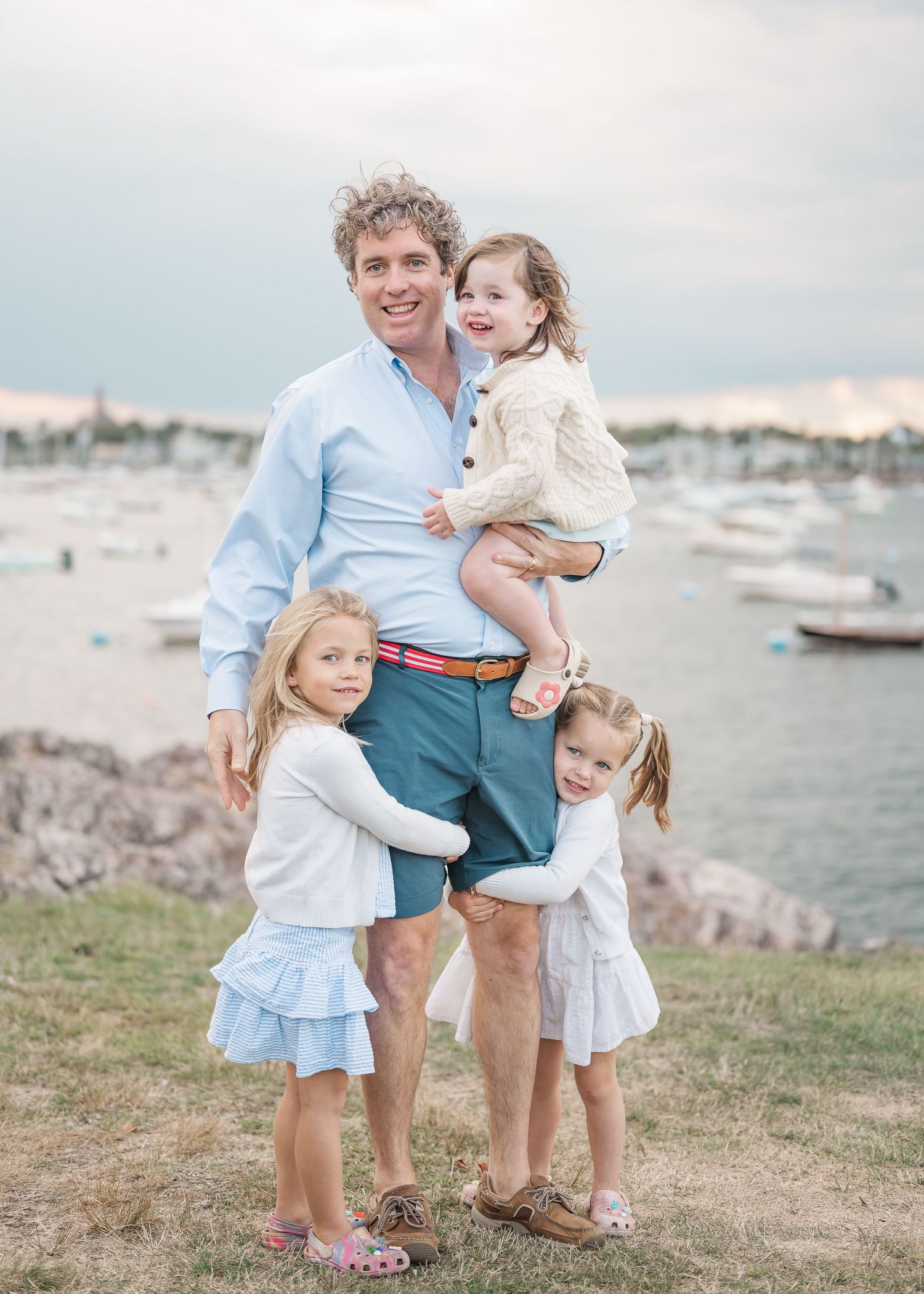 A family poses together on a grassy shore with boats and water in the background during golden hour.