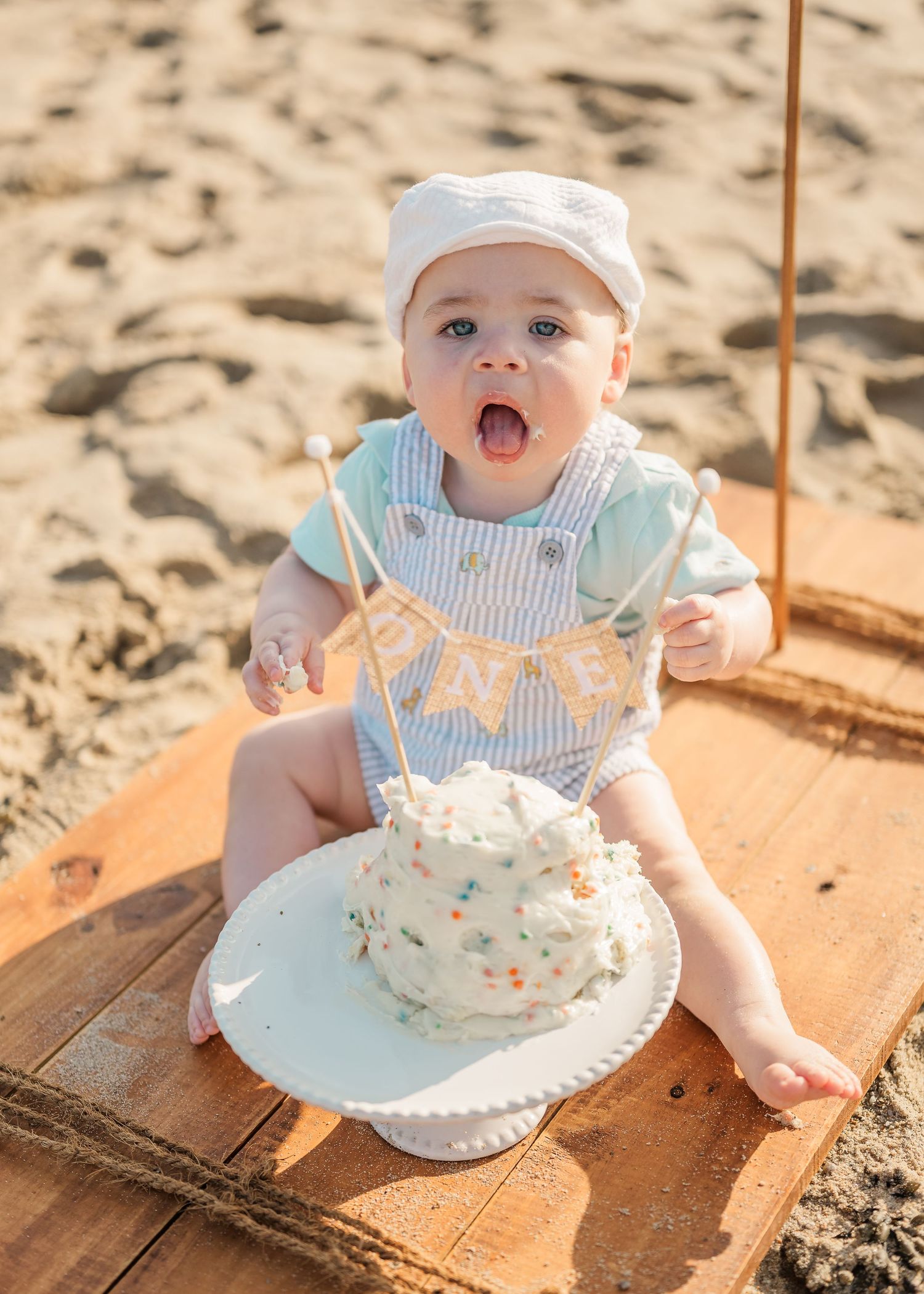Happy baby in summer outfit enjoying cake smash photo session on wooden platform at sandy beach.