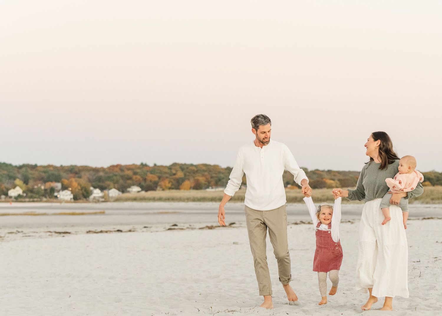 A family in light-colored clothing walks together on a sandy beach during sunset with soft pastel colors in the sky.
