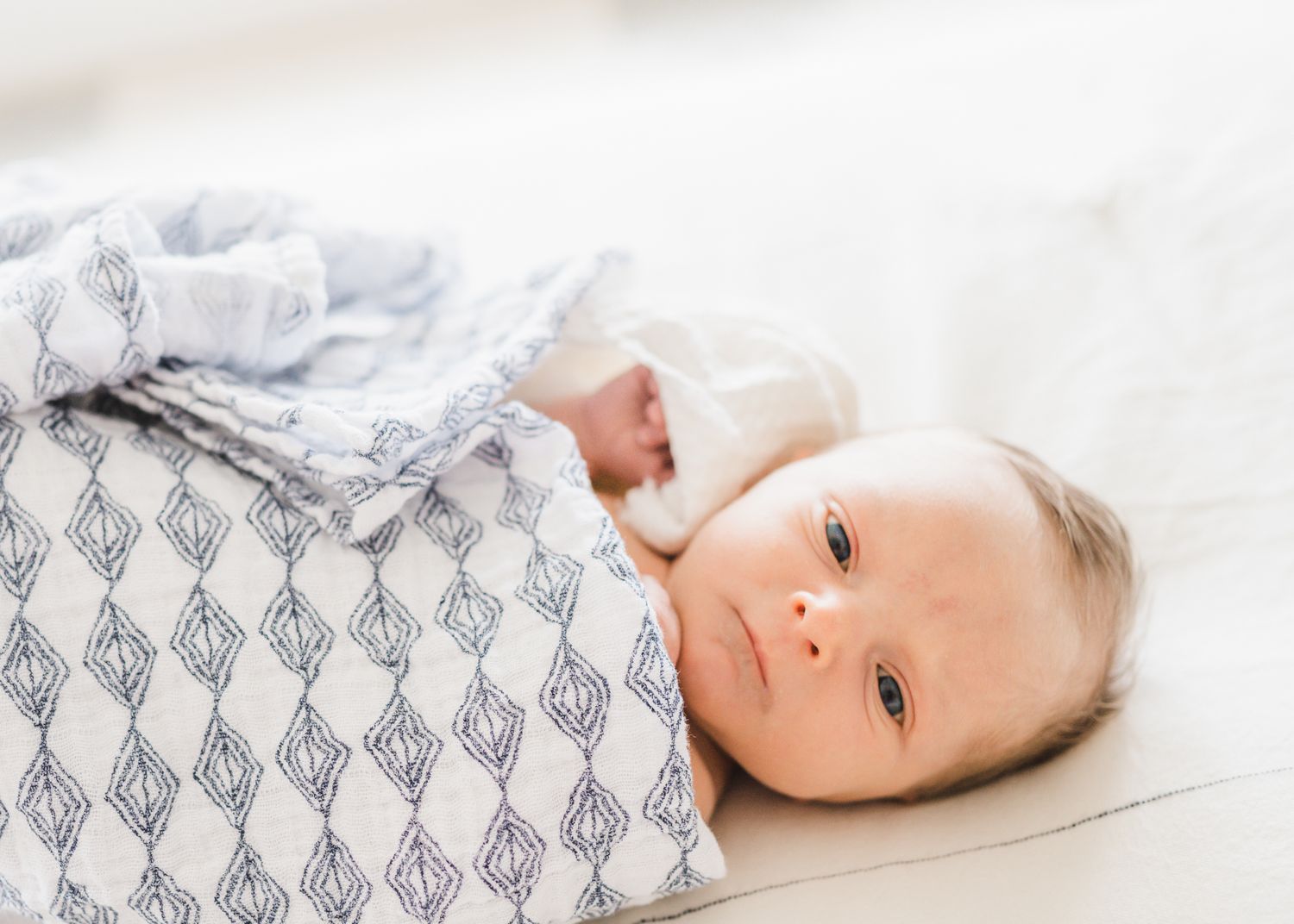 Baby lying on white surface wrapped in patterned gray and white swaddle.