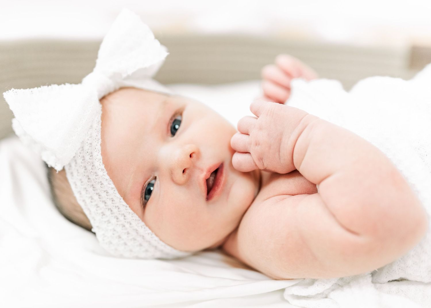 A sweet baby wearing a white headband lies contentedly on soft white bedding.