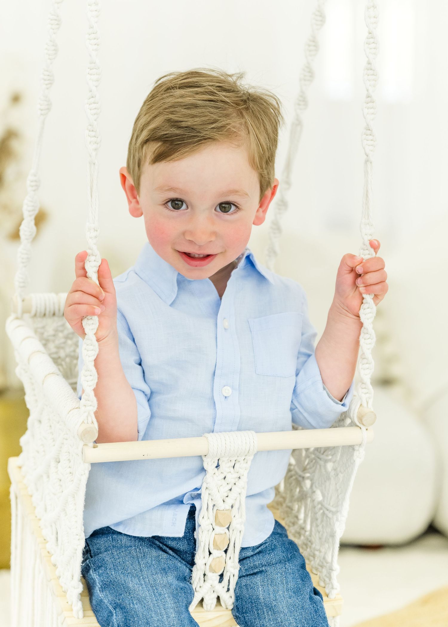 A young child in a light blue shirt and jeans sits on a white decorative swing indoors.