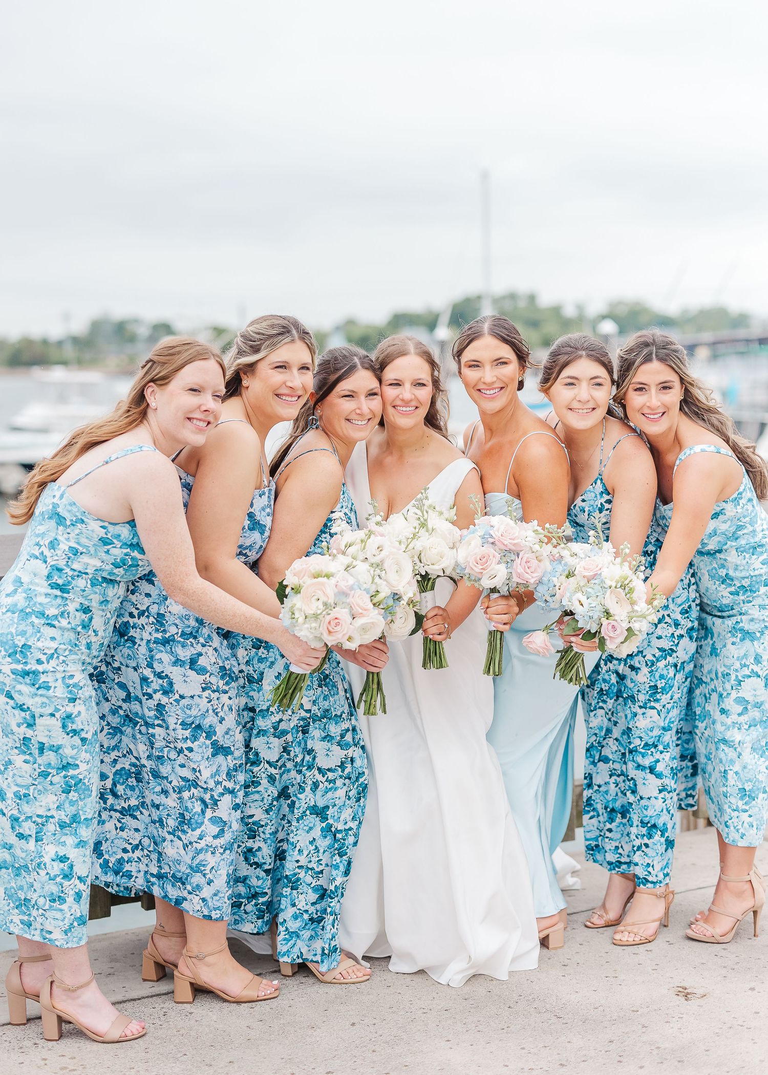 Wedding party in coordinated blue floral dresses posed together at waterfront location.
