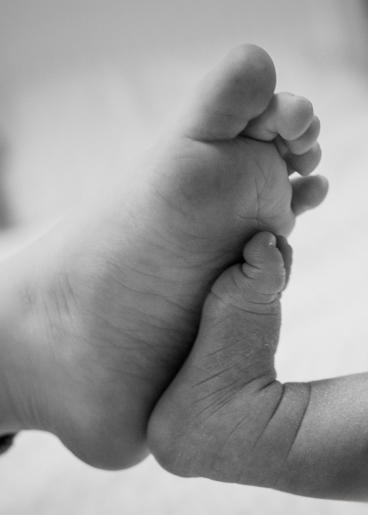 A black and white close-up photograph of a tiny baby foot next to an adult thumb in soft focus.