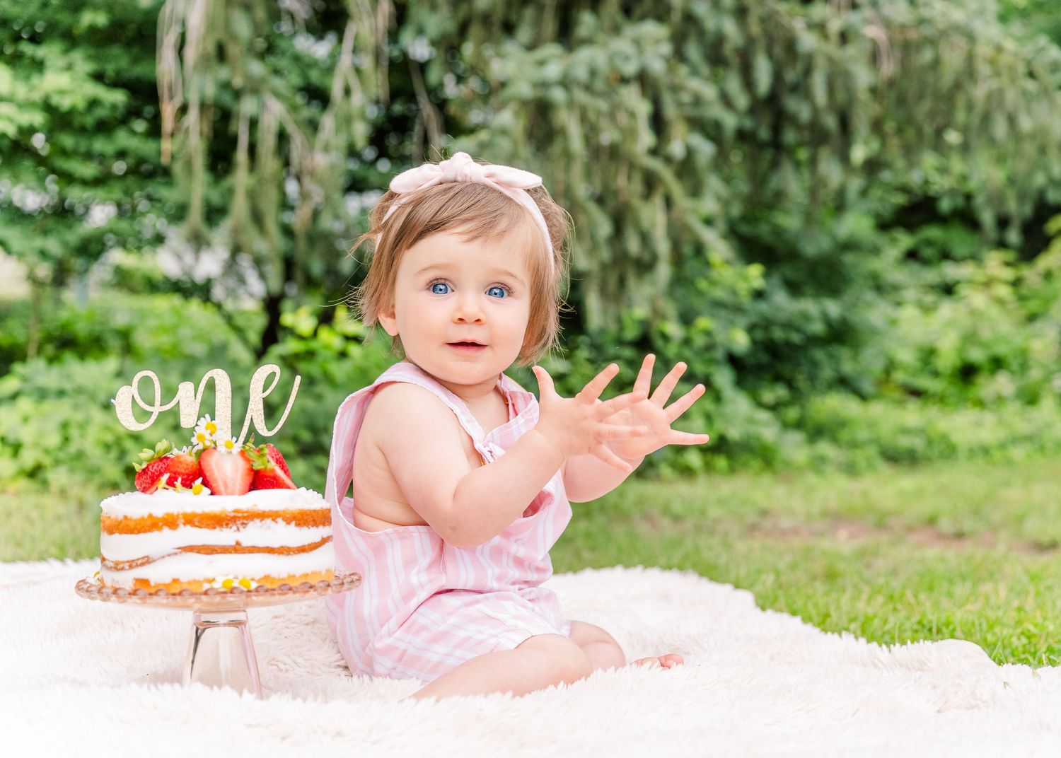 First birthday celebration with cake and pink outfit on white blanket outdoors.