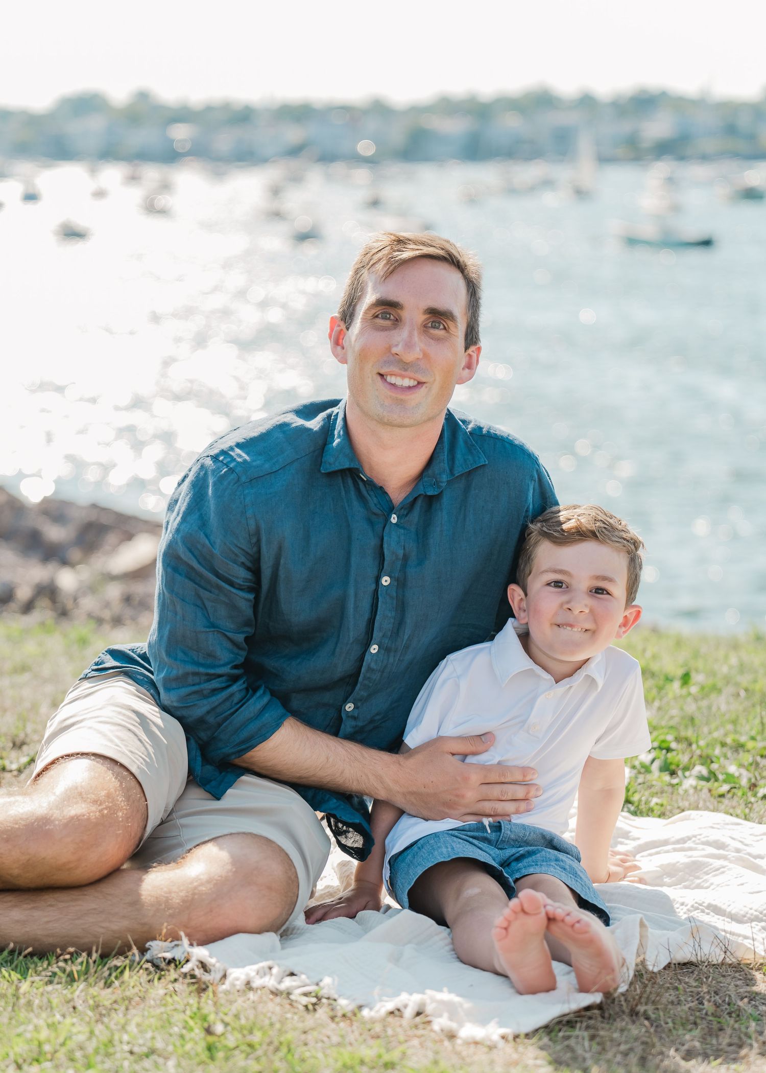 A father and son sit together on a blanket by the ocean with boats visible in the harbor behind them.