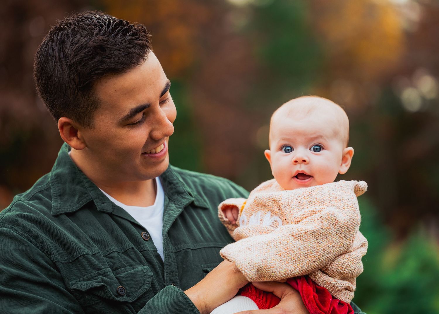 A sweet family moment captured outdoors with a small baby in a festive outfit against an autumn background.