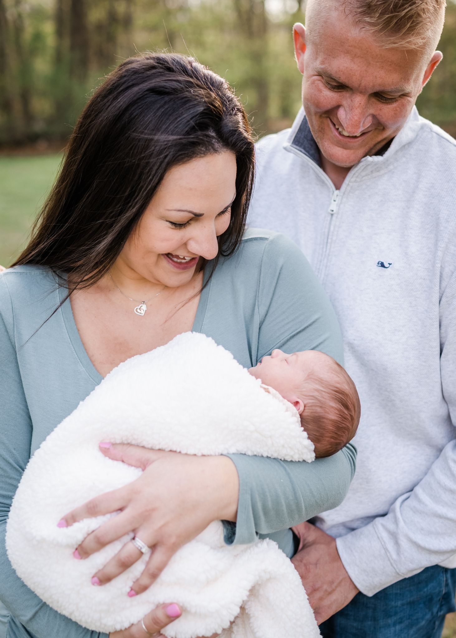 Family shares tender moment outdoors while holding newborn wrapped in white blanket.