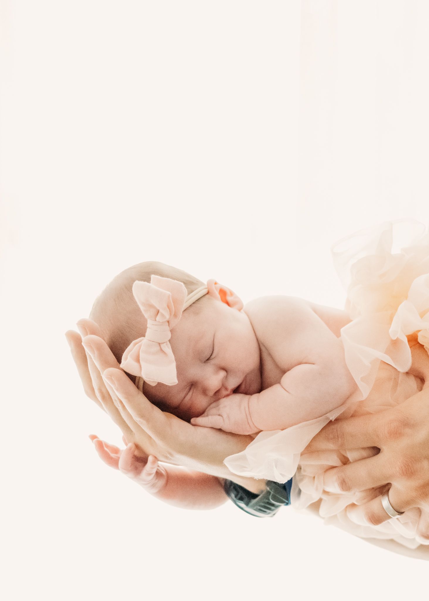 Peaceful newborn baby cradled in soft hands against light background with ethereal peach tones.
