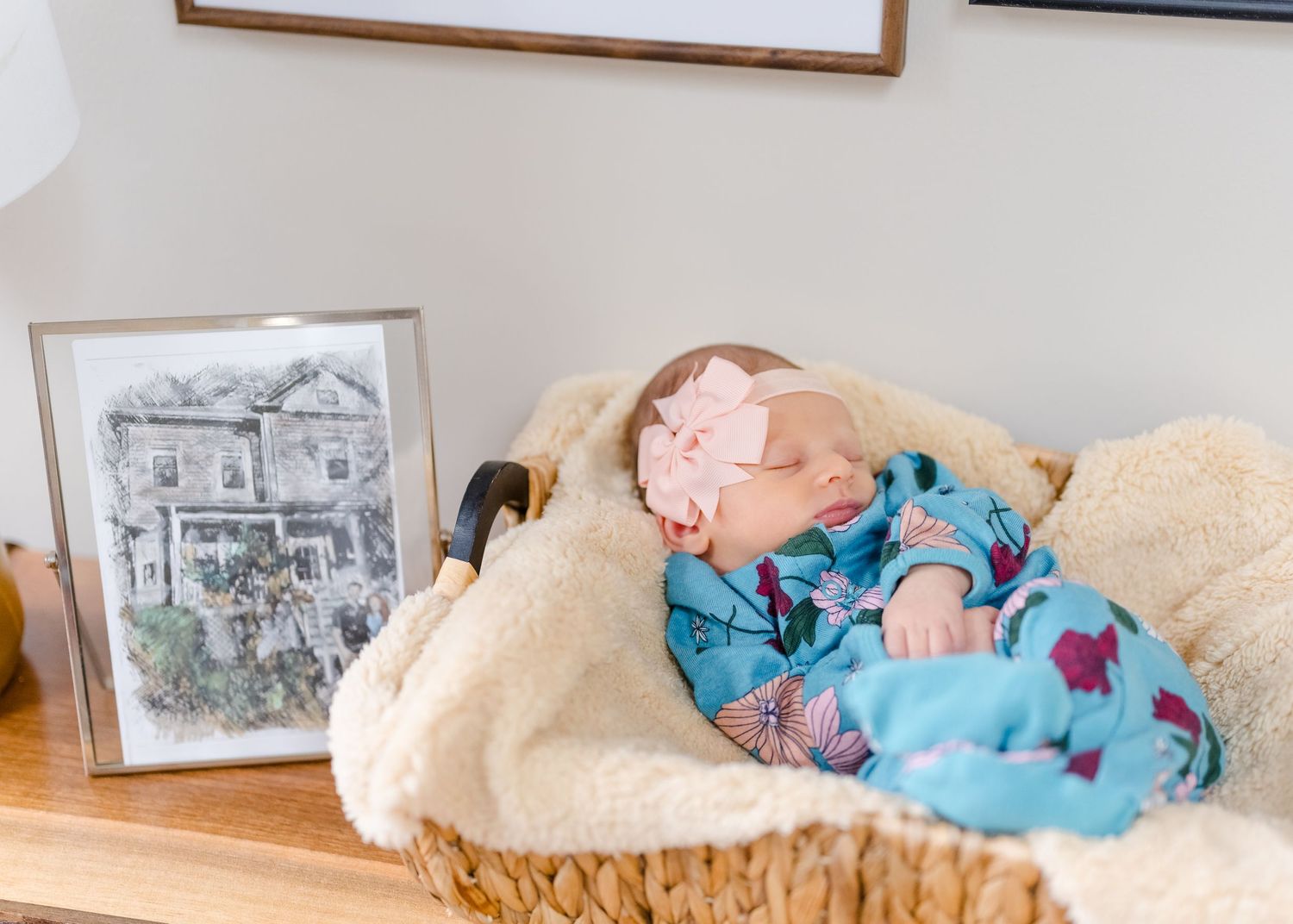 A sleeping baby wearing blue pajamas rests peacefully in a cozy basket with a framed artwork nearby.