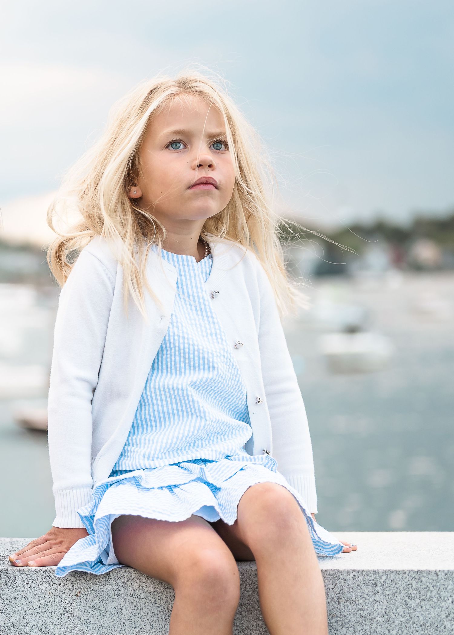 A child in a white cardigan and blue striped dress sits thoughtfully on a concrete wall near a marina on a summer day.
