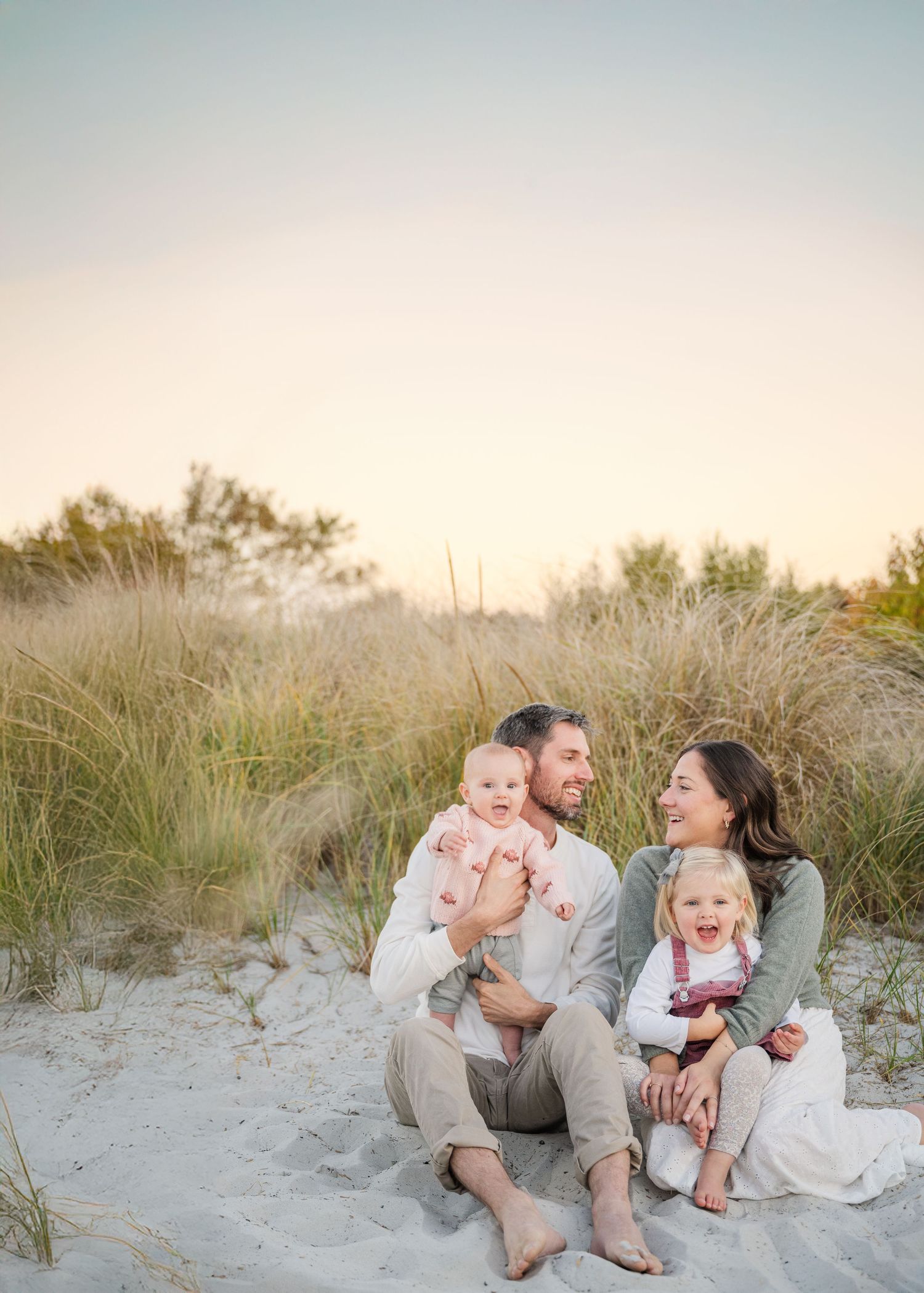 A family enjoying a playful moment together while sitting on white sandy beach dunes at sunset.